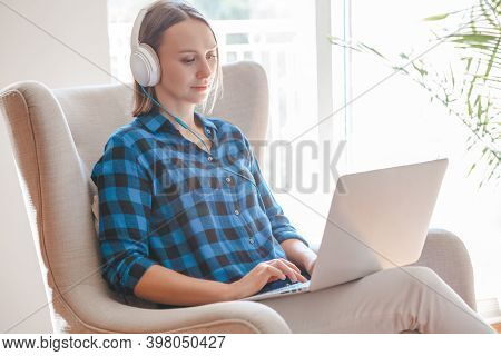 Work From Home. Middle Age Woman In Headphones Sitting In Armchair Working On Laptop Computer. Remot