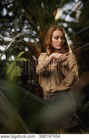 A Beautiful Girl With Natural Make-up And Red Hair Stands In The Jungle Among Exotic Plants With Sna