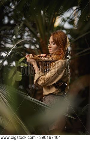 A Beautiful Girl With Natural Make-up And Red Hair Stands In The Jungle Among Exotic Plants With Sna
