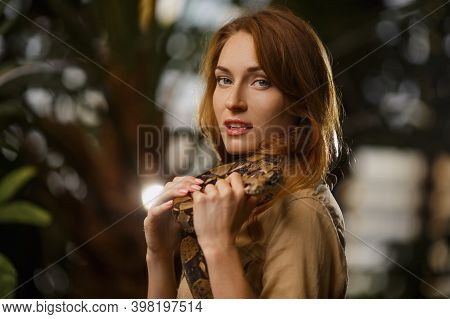 A Beautiful Girl With Natural Make-up And Red Hair Stands In The Jungle Among Exotic Plants With Sna