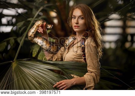 A Beautiful Girl With Natural Make-up And Red Hair Stands In The Jungle Among Exotic Plants With Sna