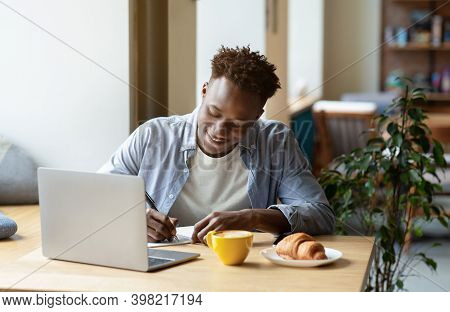 Happy Black Man Studying Remotely In Front Of Laptop, Taking Notes During Online Class Or Lecture At