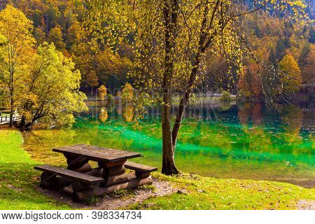 Alps, Northern Italy. Lake Fuzine. Cozy picnic corner - wooden table with benches. Yellow and orange trees are reflected in the green smooth water of the lake