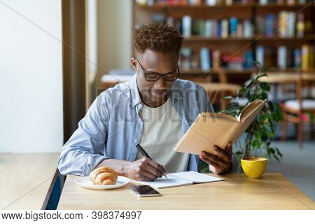 Funky Black Guy With Textbook Taking Notes In Copybook At Cozy Cafe. Positive African American Man S