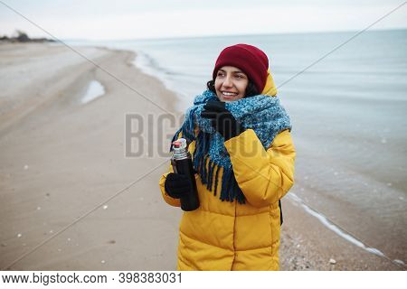 Young Woman Pours Hot Tea From A Thermos Into A Cup And Drinks On The Seaside. Female Tourist Is Hav