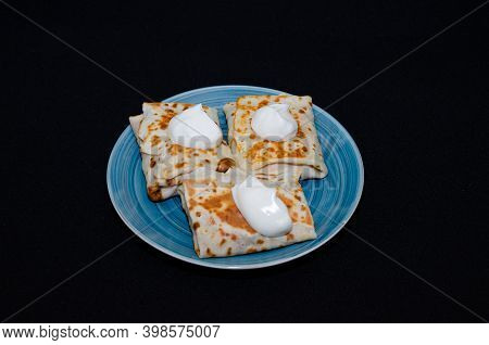Russian Thin Pancakes With Sour Cream And Filling On A Blue Plate, On A Black Background