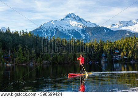 Adventurous Girl Paddle Boarding On Levette Lake With Famous Tantalus Mountain Range In The Backgrou