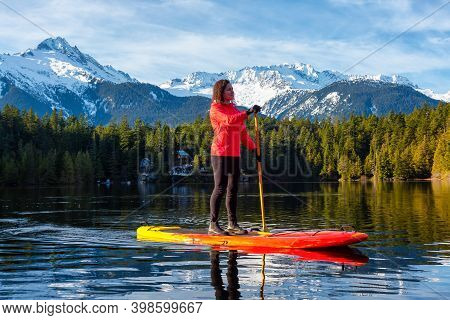 Adventurous Girl Paddle Boarding On Levette Lake With Famous Tantalus Mountain Range In The Backgrou