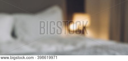 Cozy Hotel White Bed In Room And Reading Lamp Blurred Focus Background. Blur Bedroom With Glowing La