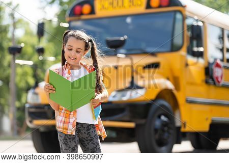 Adorable Little Schoolgirl Near School Bus And Looking At Camera