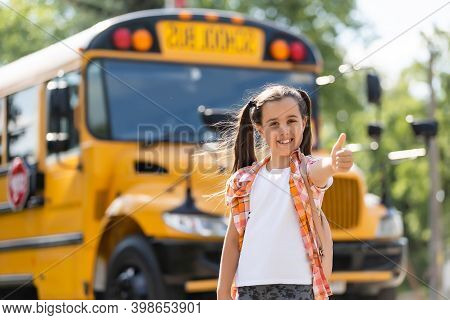 Adorable Little Schoolgirl Near School Bus And Looking At Camera