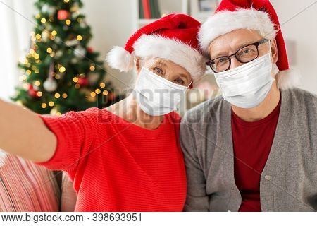 christmas, holidays and health concept - happy smiling senior couple wearing face protective medical mask for protection from virus disease in santa hats taking selfie at home