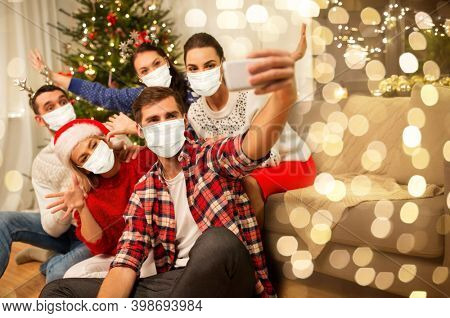 health, people and winter holidays concept - happy friends with taking selfie by smartphone wearing face protective medical mask for protection from virus disease at home on christmas
