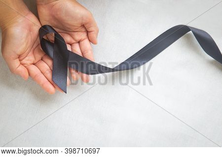 Hand Holding Black Ribbon On White Fabric Background With Copy Space, Symbol Of Skin Cancer Awarenes