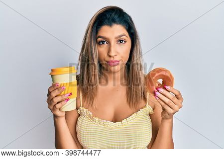 Beautiful brunette woman eating doughnut and drinking coffee relaxed with serious expression on face. simple and natural looking at the camera. 