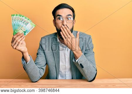Young hispanic man holding russian ruble banknotes sitting on the table covering mouth with hand, shocked and afraid for mistake. surprised expression 