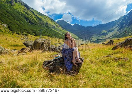Sensual Pretty Girl In Long Blue White Dress Sits On A Rock And Dreaming Against The High Mountains 
