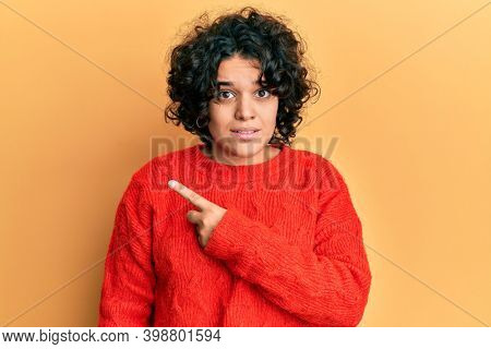 Young hispanic woman with curly hair wearing casual winter sweater pointing aside worried and nervous with forefinger, concerned and surprised expression 
