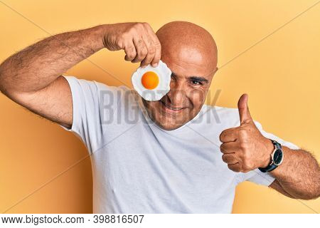 Mature middle east man holding fried egg smiling happy and positive, thumb up doing excellent and approval sign 