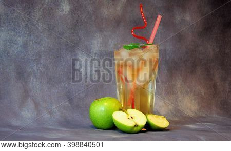 A Tall Glass Of Pale Yellow Fruit Juice With Ice And Straws Stands Against A Gray Background Next To