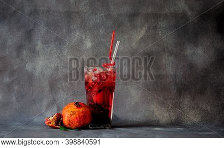 A Tall Glass Of A Red Drink With Ice And Straws Stands On A Gray Background Next To Ripe Pomegranate