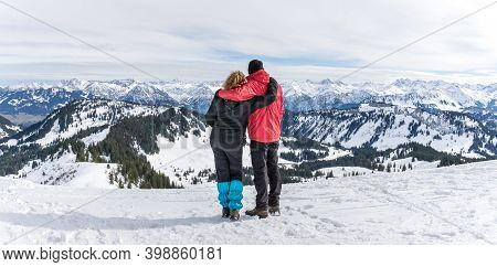 Senior Couple Is Hiking In Alpine Snow Winter Mountains Enjoying Panorama View. Allgau, Bavaria, Ger