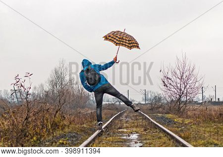 The Man Walks On The Railway Trying To Keep His Balance. He Has A Backpack And An Umbrella In His Ha
