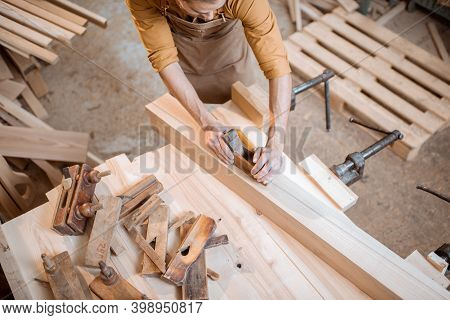 Handsome Carpenter Working With A Wood, Planing A Bar With A Plane On The Worbench In The Carpentry 
