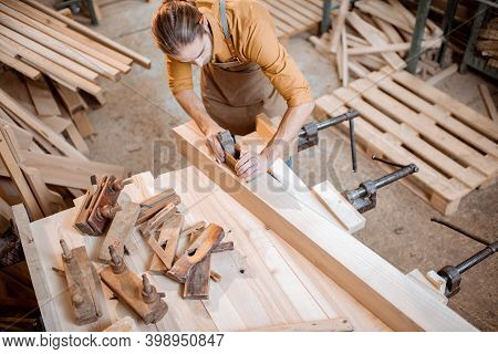 Handsome Carpenter Working With A Wood, Planing A Bar With A Plane On The Worbench In The Carpentry 