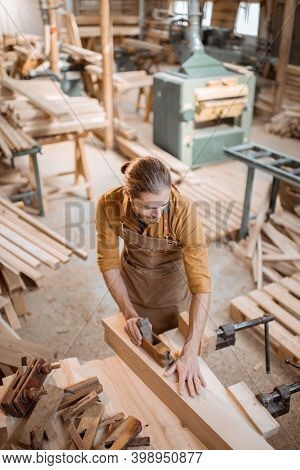 Handsome Carpenter Working With A Wood, Planing A Bar With A Plane On The Worbench In The Carpentry 