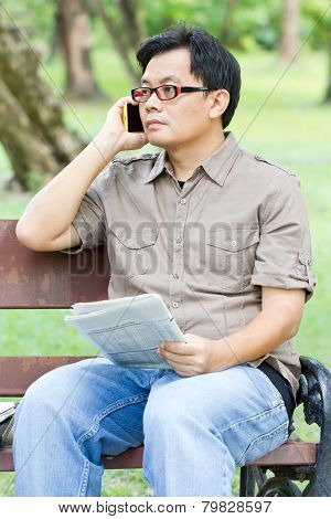 Asiam Man Talking On Phone With Newspaper In Hand.