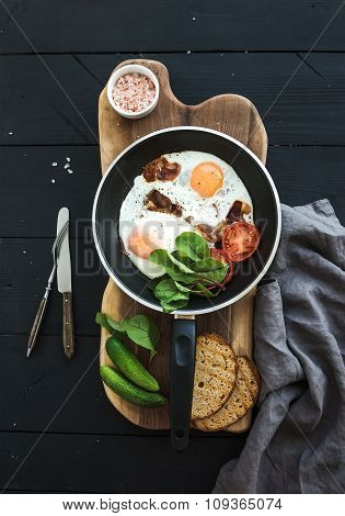 Pan of fried eggs, bacon, tomatoes with bread, mangold and cucumbers on rustic wooden serving board
