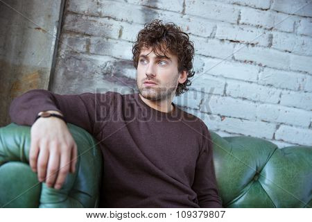 Pensive attractive thoughtful curly young man in brown sweetshirt thinking on green leather sofa alone