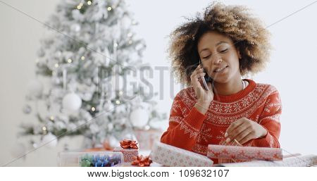 Young African woman wrapping gifts at Christmas sitting at a table in front of the Xmas tree chatting on her mobile phone  natural pre-occupied pose.