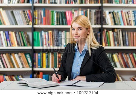 Smiling Young Girl With Blonde Hair  Sitting At A Desk In The Library With An Open Note Book Making