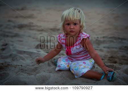 Small Blonde Girl In Colorful Sits On Beach Sand Looks Forward