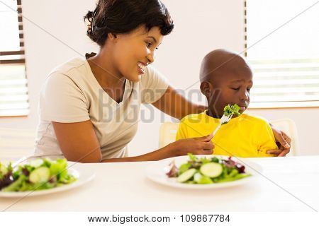 pretty african mother trying to feed her little son who hates eating green salad