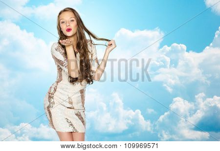 people, style, holidays, hairstyle and fashion concept - happy young woman or teen girl in fancy dress with sequins and long wavy hair sending blow kiss over blue sky and clouds background