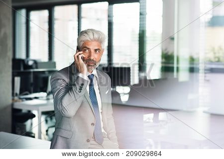 Mature businessman in gray suit in the office standing at the window holding smartphone making phone call. Shot through glass.
