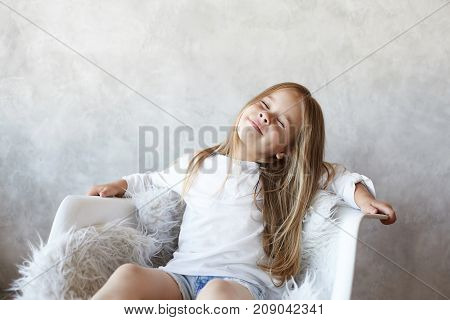 Indoor shot of playful cute little girl with fair hair wearing beautiful white blouse sitting in armchair and throwing her head back closing eyes fantasizing and dreaming about something pleasant