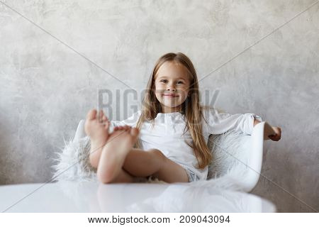 Cheerful pretty little girl with charming smile having relaxed and carefree look spending weekend morning at home sitting on white armchair resting bare feet on table ready to watch cartoons