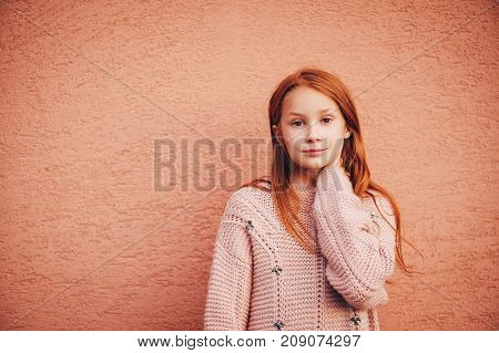 Outdoor portrait of adorable red-haired preteen girl wearing soft pink pullover kid model posing against beige wall background. Fashion for young teens