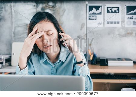 Stressed Asian creative designer woman cover her face with hand and feel upset while talk on mobile phone with customer in front of laptop computer on desk at office Stress office lifestyle concept.