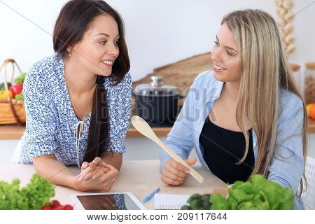 Two young happy women are making online shopping by tablet computer and credit card. Friends are going to cook in the kitchen, they searching internet for a new recipe