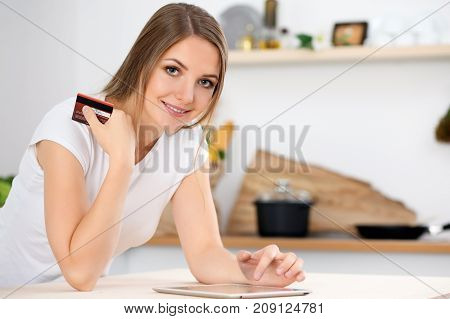 Young woman is making online shopping by tablet computer and credit card. Housewife found new recipe for cooking in a kitchen.