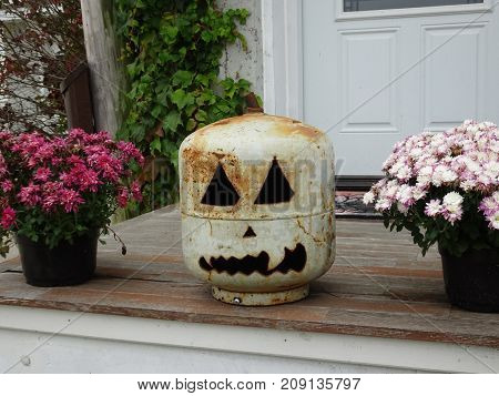 Jack o lantern created from repurposed white rusty gas can sits between two colorful mum plants on wooden steps in a fall display.