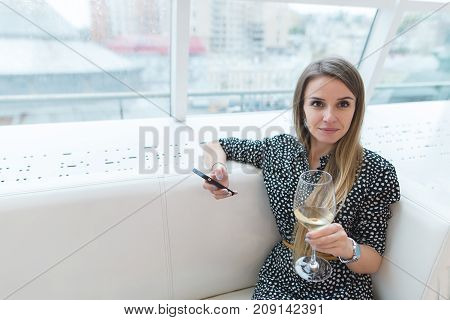 Beautiful woman sitting on a sofa in a stylish restaurant with a glass of wine and a smartphone in her hands