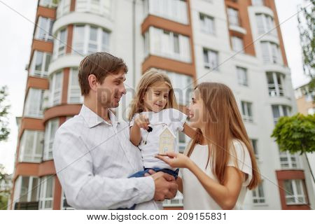 Cheerful parents holding their little daughter and keys to their new home in apartment building on the back. happy family outdoors.Loving family embracing posing in front of a new apartment building