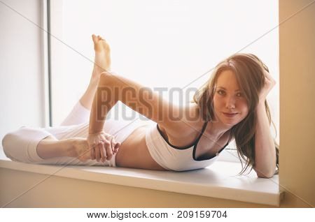Beautiful sporty fit yogini woman practices yoga asana Ardha Bhekasana - Half Frog pose in a window sill.