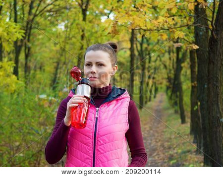 Beautiful fitness sport girl in autumn park in sportswear drinks water or isotonic drink from a sports bottle